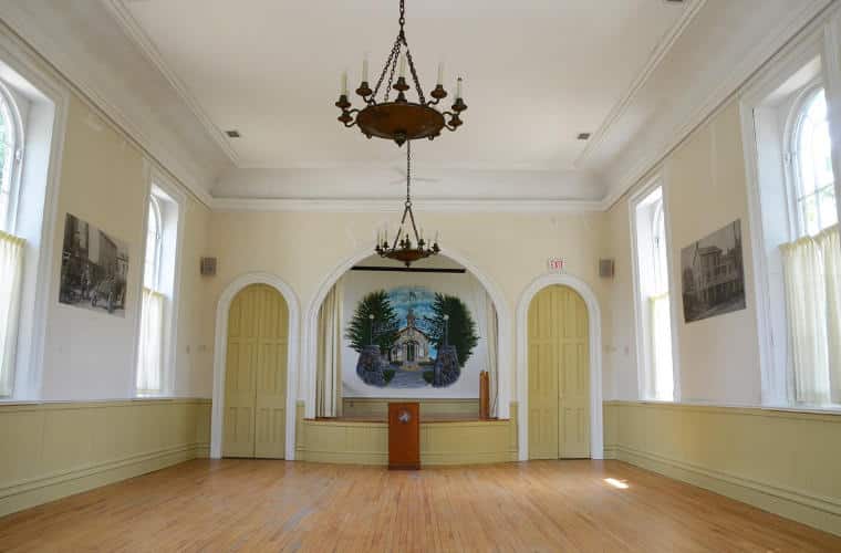 A large white room with curtained windows, hardwood floor, and podium at the far end.