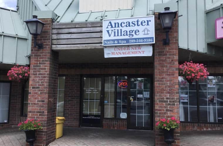 A building with a green roof, wall window first floor, and red brick pillars in front of the entrance with a sign for Ancaster Village Nails and Spa between them.