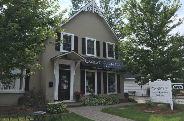 A grey house with large peaked roof and a sign for Caniche French Bakery on the front lawn.