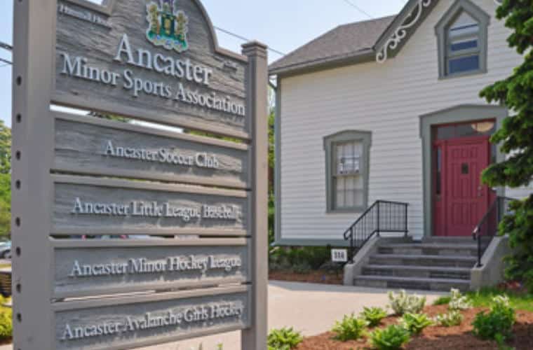 A building with white siding and a red door with a pylon sign on the front lawn with numerous sports clubs listed on it.