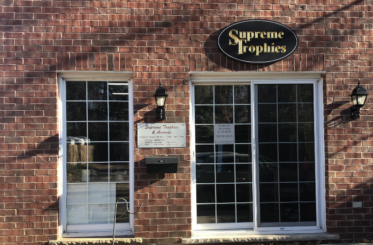 The front of a red brick building with a sign for Supreme Trophies Awards above the patio door.