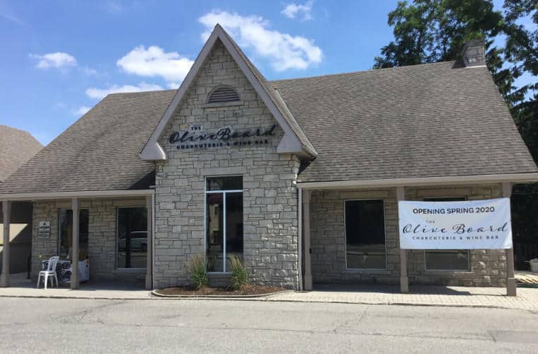 A rough hewn grey briock building with large peaked roof and a sign for The Olive Board above the entrance.